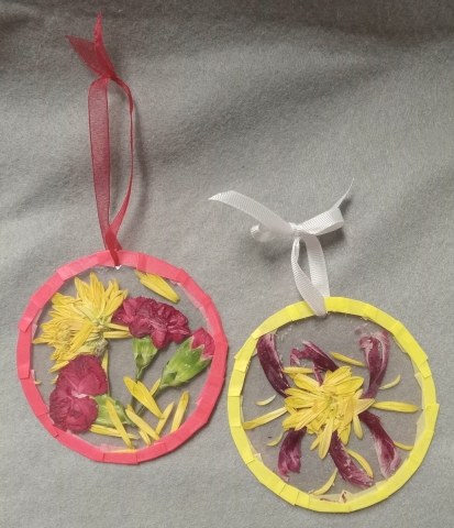 Two circles of clear plastic sun catchers with pressed red, yellow and purple flowers. One with a red border and red ribbon, one with yellow border and white ribbon.