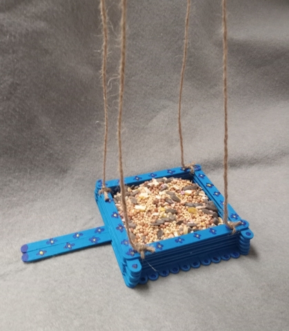 Popsicle sticks formed to create a basket style bird feeder, painted blue with purple flowers. Brown twine used as the string to hang feeder.
