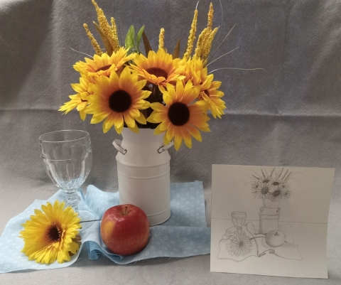 White vase of sunflowers with single sunflower on table with red apple and glass, sitting on top of a blue cloth. Drawing of still life arrangement next to flower vase.  
