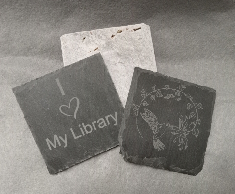 Three coasters, one white one, two black slate with engravings on them. One has "I heart my library" the other with a hummingbird and flowers.