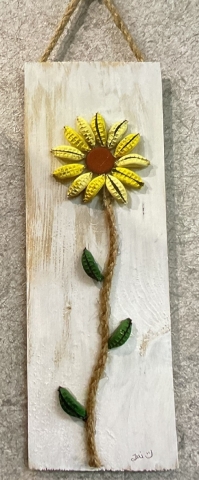 A light colored wood panel hangs against a beige wall with twisted rope. On the board is a sunflower composed of yellow bottle caps that have been crimped together, a rope stem, and green leaves made from green bottle caps crimped together. bottl