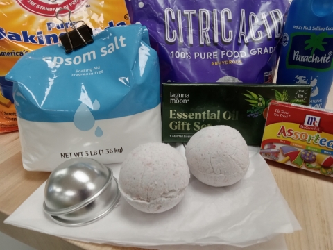Two white bath bombs shown with bags of baking soda, citric acid, corn starch and coconut oil, fragrance, food coloring and two silver molds.
