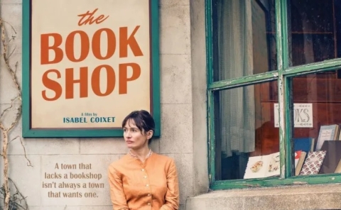 Young woman (Emily Mortimer) in an orange blouse leans against a quarried stone bookshop storefront with the bookshop display window on her right and a large sign reading "the Book Shop" above her to the left